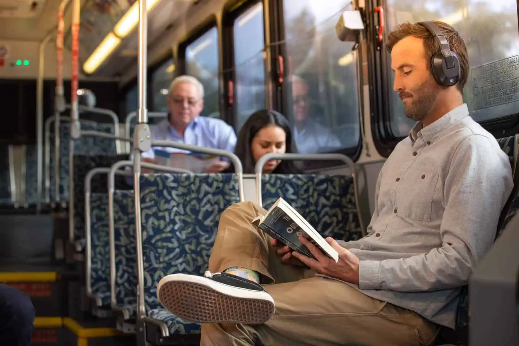 Man on bus sitting with book and headphones legs crossed with two others warm color